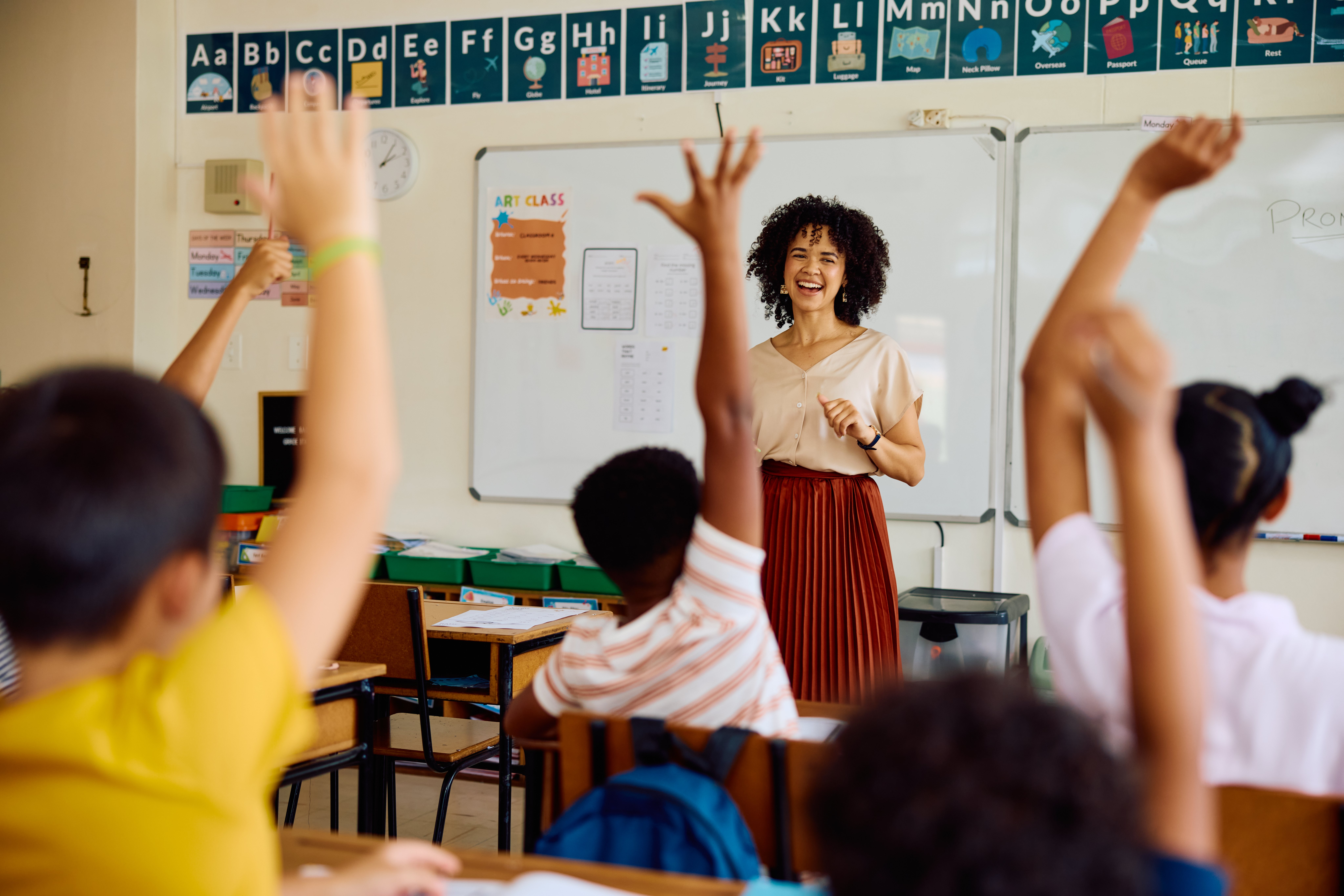  Elementary school teacher smiling and asking question, children with hands raised