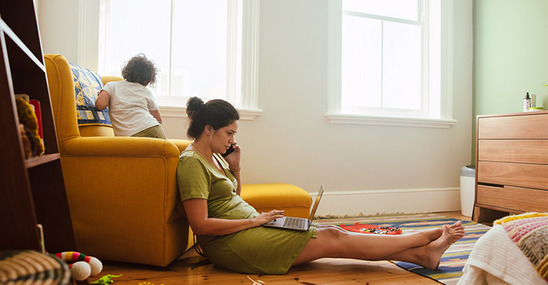 busy mom seated on the floor on the phone and computer, with child playing nearby