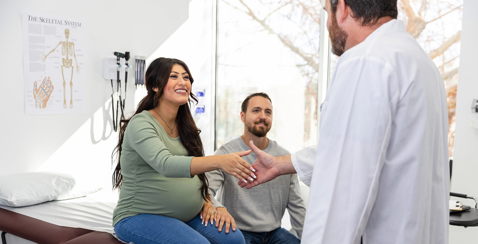 smiling pregnant woman in exam room with her partner, shaking hands with her provider