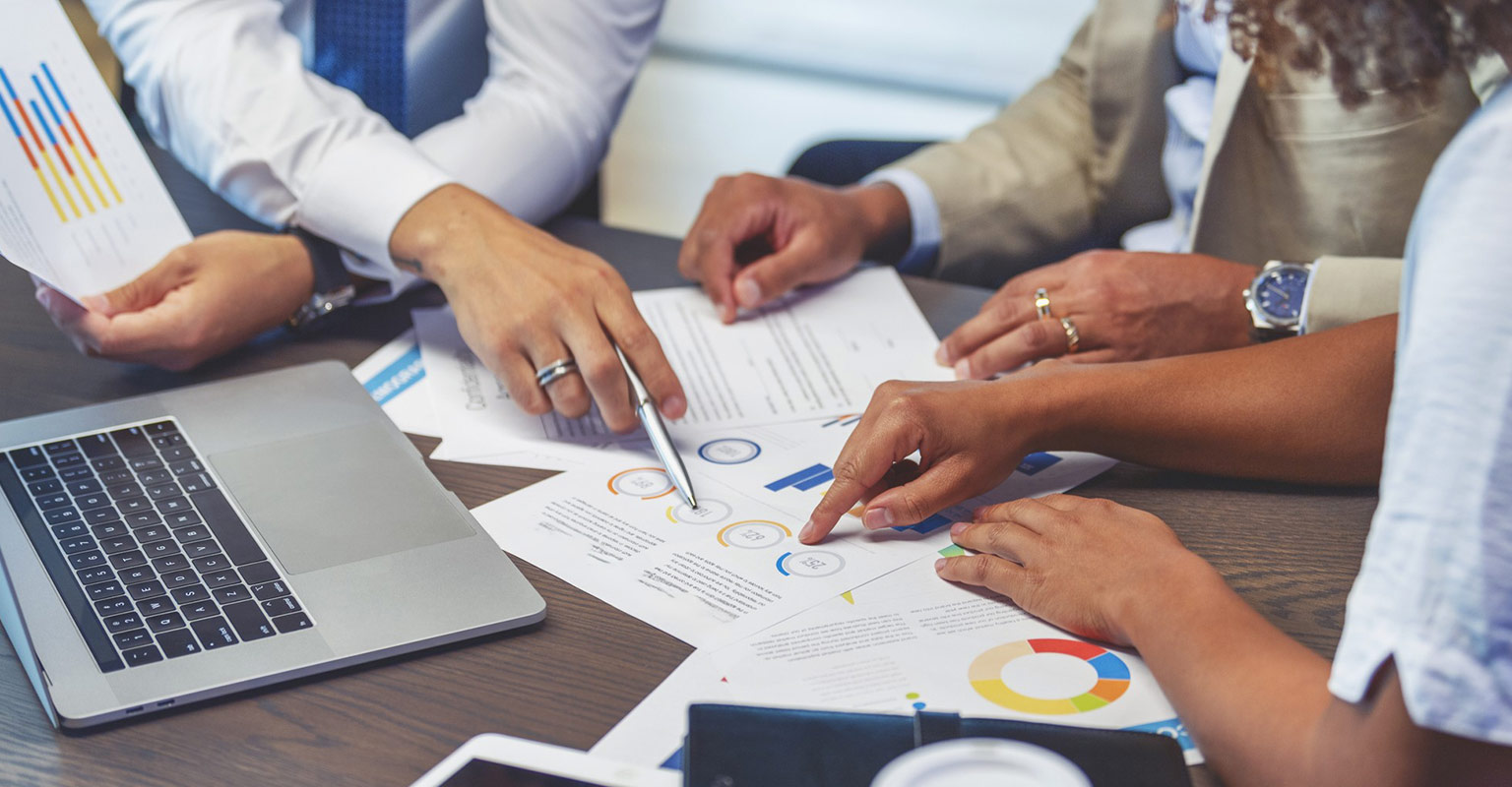 closeup image of table with paperwork, laptop, and colleagues working
